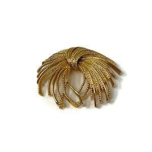 May include: A gold-toned brooch with a circular design. The brooch is made of multiple strands of metal that are twisted and woven together to create a radial pattern.
