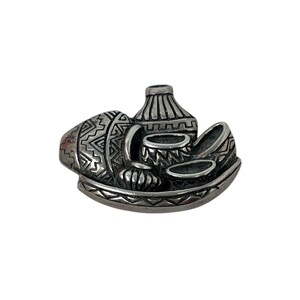 May include: A silver-toned brooch featuring a still life of pottery. The brooch depicts various vessels, including a vase, bowls, and a small pot, all arranged on a decorative base. The pottery has detailed patterns.