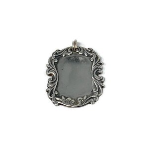 May include: A silver pendant with an ornate, scrolled frame. The pendant is blank and ready to be engraved.