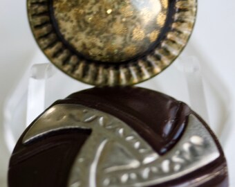 May include: Two decorative buttons with a silver and brown design. The top button has a round, silver-colored center with a textured surface and a brown outer ring with a raised edge. The bottom button is brown with a silver, geometric design.