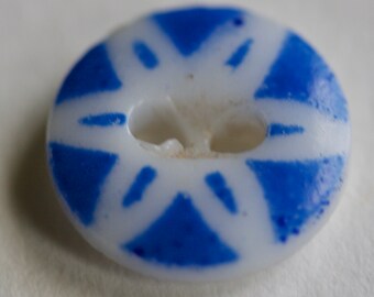 May include: A round, white button with a blue geometric design. The button has two holes for sewing and a pattern of blue triangles and lines radiating from the center. The button is likely made of plastic or glass.