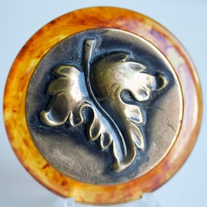 Puede incluir: Un botón redondo de color ámbar con un diseño de hoja en relieve de color dorado. El botón tiene una superficie lisa y brillante.