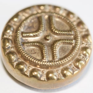 Könnte beinhalten: Nahaufnahme eines Messingknopfs mit Kreuzmuster. Der Knopf hat ein erhabenes Kreuzdesign in einem kreisförmigen Rahmen, umgeben von einem Perlenrand. Der Knopf hat ein Vintage-Aussehen und ein metallisches Finish.
