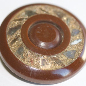 May include: A brown plastic button with a gold-colored design. The button has a raised center and a decorative border.