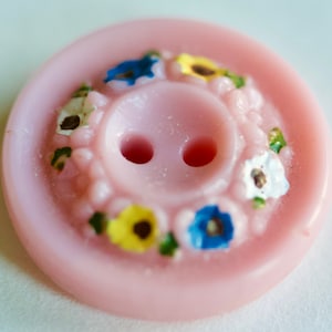 May include: A light pink button with two holes and a floral design. The button has a raised center with two holes and is encircled by small, colorful flowers in blue, yellow, and white.