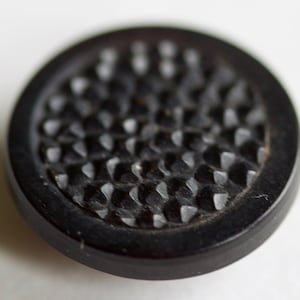 Puede incluir: Un botón negro con un diseño texturizado en relieve. El botón tiene un borde negro liso.