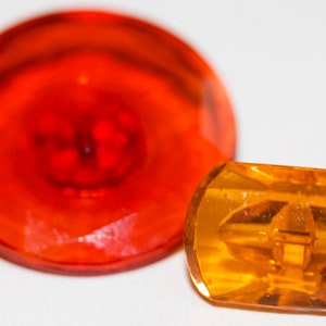 May include: Two decorative glass buttons. One is a large, round, faceted button in a translucent orange color with a red center. The other is a smaller, rectangular button in a translucent amber color.
