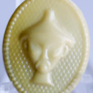 Puede incluir: Un botón de plástico blanco con una imagen en relieve detallada de un extraterrestre con un sombrero. El botón tiene un borde circular texturizado.