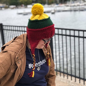 May include: A red, green, and yellow knitted hat with a yellow pom-pom on top. The hat has long ear flaps with yellow and red tassels. The hat is worn by a person wearing a blue shirt with the text "#blessed" on it.