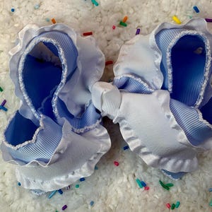 May include: Two light blue and white hair bows. The bows are made of layered ribbons with a ruffled edge. The center of the bows are tied with a white ribbon. The bows are on a white, textured surface with colorful sprinkles.