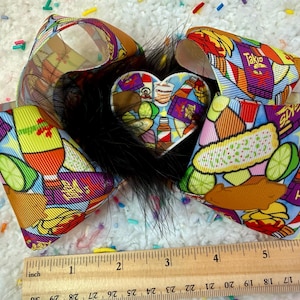 May include: A colorful hair bow featuring a heart-shaped center and ribbon with food graphics. The bow includes black feathers and a ruler for scale. The ribbon design includes images of tacos, limes, and bottles.