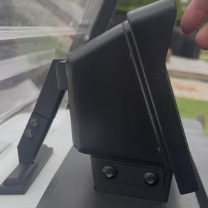 May include: A black plastic monitor housing with an integrated sun visor, mounted on a black bracket with two visible screws. The unit features a sleek, angled design and is attached to a support arm that connects to a flat, horizontal surface.