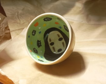 May include: A small ceramic bowl with a green glaze and a painted design of a white No Face character from the movie Spirited Away. The bowl is decorated with black and yellow dots and a few small black creatures.