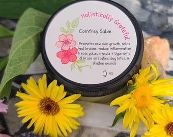 May include: A small jar of Comfrey Salve with a white label that reads "Holistically Grateful Comfrey Salve" and lists the benefits of the salve. The jar is sitting on a surface with flowers.