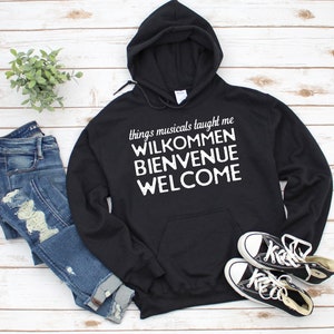 Op de afbeelding: Zwart hoodie met de tekst "things musicals taught me" en de woorden "Willkommen", "Bienvenue" en "Welcome" in witte letters.