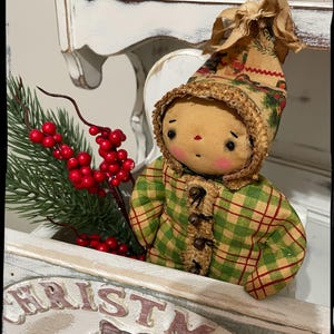 May include: A handmade rag doll wearing a green and red plaid coat and a brown hat with a floral print. The doll is sitting in a white wooden box with the word "CHRISTMAS" painted on it. The doll is holding a sprig of green pine with red berries.