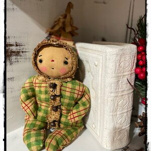 May include: A handmade rag doll wearing a green and red plaid outfit with a brown fur trim. The doll has a round face with painted features and is sitting on a white shelf next to a white book.