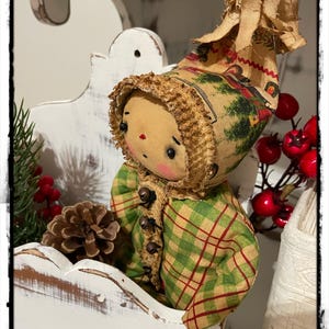 May include: A handmade rag doll wearing a green and red plaid coat and a brown and white hat with a winter scene. The doll is sitting in a white wooden shelf with a pine bough and pine cone behind it.