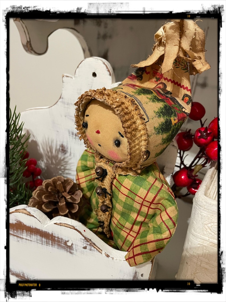 May include: A handmade rag doll wearing a green and red plaid coat and a brown and white hat with a winter scene. The doll is sitting in a white wooden shelf with a pine bough and pine cone behind it.