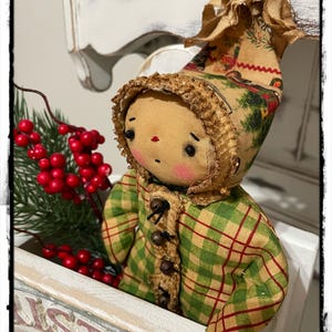 May include: A handmade rag doll wearing a green and red plaid coat with a brown fur trim. The doll is wearing a brown hat with a white trim and has a red nose and black eyes. The doll is sitting in a white wooden box with the word "Christmas" written on it.