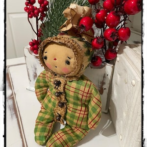 May include: A handmade rag doll wearing a green and red plaid outfit with brown buttons. The doll has a brown yarn wig and a brown felt face with painted features. The doll is sitting on a white surface with a sprig of red berries and greenery behind it.