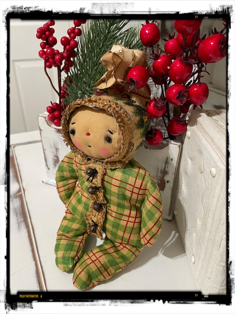 May include: A handmade rag doll wearing a green and red plaid outfit with brown buttons. The doll has a brown yarn wig and a brown felt face with painted features. The doll is sitting on a white surface with a sprig of red berries and greenery behind it.