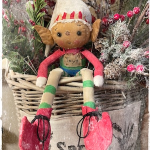 Pode incluir: Um boneco de duende feito à mão com um chapéu listrado vermelho e branco, pernas listradas verde e branco e sapatos vermelhos. O duende está sentado numa cesta de vime com um saco de estopa que diz "Oficina do Papai Noel".
