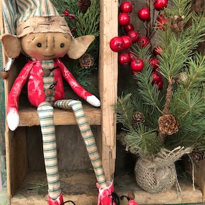 Pode incluir: Uma boneca de elfo feita &agrave; m&atilde;o com uma camisa listrada vermelha e branca, cal&ccedil;as listradas verdes e brancas e sapatos vermelhos. O elfo est&aacute; sentado em uma prateleira de madeira com um ramo de pinheiro verde e bagas vermelhas no fundo.