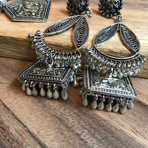 Puede incluir: Un par de pendientes de plata con dise&ntilde;os intrincados y un colgante de forma cuadrada en la parte inferior. Los pendientes tienen una cadena de plata con peque&ntilde;as cuentas de plata que cuelgan de la parte inferior.