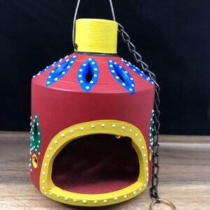 Könnte beinhalten: Eine rote, hängende Terrakotta-Lampe mit gelben, blauen und grünen bemalten Designs. Die Lampe hat eine Kette zum Aufhängen und eine runde Öffnung für eine Kerze oder ein Teelicht.