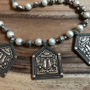 Puede incluir: Un collar de plata con cuentas de perlas blancas y tres colgantes de plata con dise&ntilde;os ornamentados. Los colgantes tienen un dise&ntilde;o en relieve de una figura con un sombrero y un motivo floral.