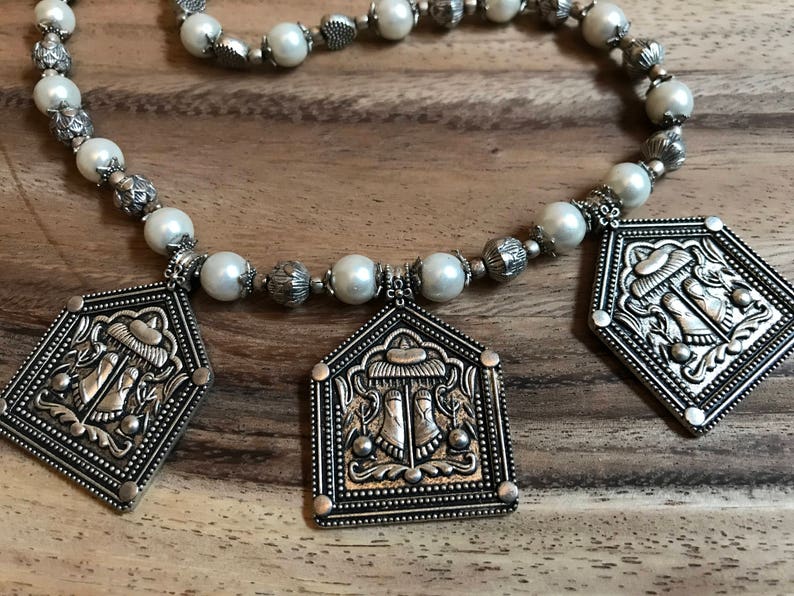 Puede incluir: Un collar de plata con cuentas de perlas blancas y tres colgantes de plata con dise&ntilde;os ornamentados. Los colgantes tienen un dise&ntilde;o en relieve de una figura con un sombrero y un motivo floral.