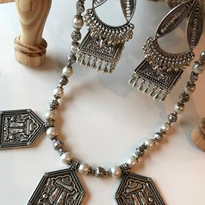 Puede incluir: Un collar de plata con cuentas de perlas blancas y tres colgantes de plata. Los colgantes tienen forma de casa con dise&ntilde;os intrincados.