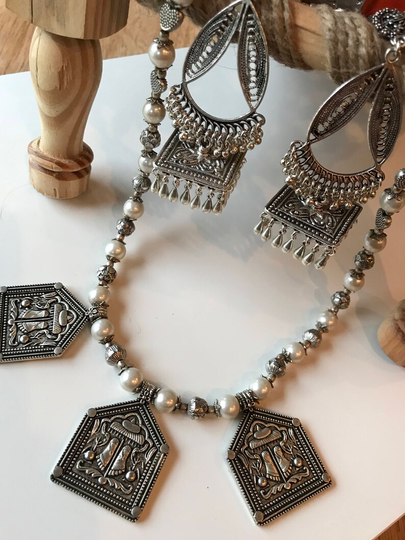 Puede incluir: Un collar de plata con cuentas de perlas blancas y tres colgantes de plata. Los colgantes tienen forma de casa con dise&ntilde;os intrincados.