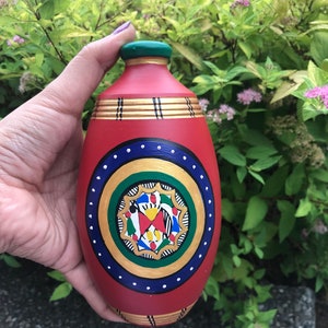 Może przedstawiać: Ręcznie malowana ceramiczna butelka w kolorze czerwonym z zielonym korkiem. Butelka ma okrągły wzór w kolorze złotym i niebieskim z wielokolorowym wzorem kwiatowym w środku.