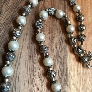 Puede incluir: Un collar de perlas blancas y plata con cuentas de plata adornadas. El collar est&aacute; hecho de perlas blancas y cuentas de plata con dise&ntilde;os intrincados.