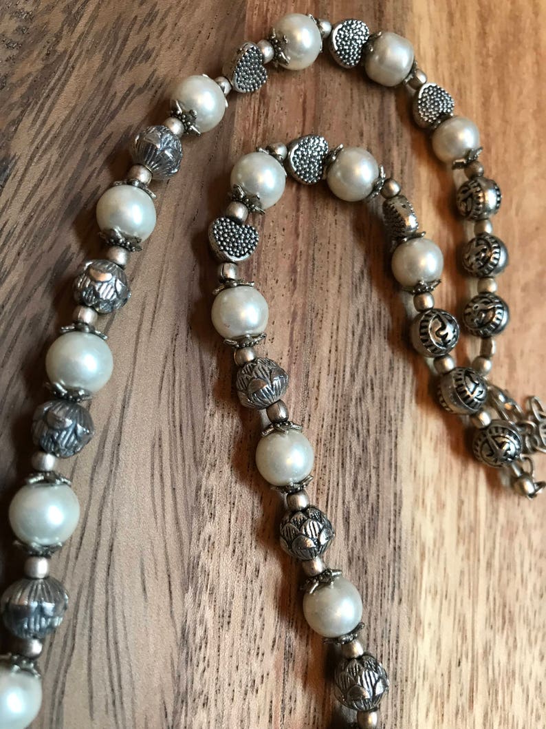 Puede incluir: Un collar de perlas blancas y plata con cuentas de plata adornadas. El collar est&aacute; hecho de perlas blancas y cuentas de plata con dise&ntilde;os intrincados.