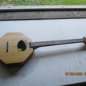 May include: A wooden, octagonal-shaped stringed instrument with a long neck and fretboard. The sound hole is decorated with a floral design. The instrument has a light brown body and a dark brown neck.