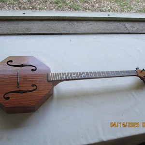 May include: A wooden octagonal guitar with a brown finish and a black fretboard. The guitar has a unique sound hole design with two f-holes.