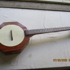 May include: A wooden banjo with an octagonal body and a long neck. The banjo has a light-colored circular soundboard and dark wood detailing. The headstock features tuning pegs. The date and time stamp "07/30/2025 09:33" is visible in the bottom right corner.