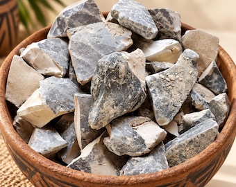 May include: A round, brown, patterned wooden bowl filled with numerous irregular, jagged chunks of grey and off-white stone. The stones have a rough, textured appearance with varying shades of charcoal, slate grey, and cream, piled high in the bowl.