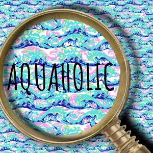 May include: A magnifying glass with a metallic frame is positioned over a repeating pattern of stylized ocean waves in shades of turquoise, light blue, dark blue, and pink. The word AQUAHOLIC is printed in black capital letters across the center.