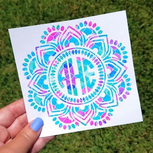 May include: A white sticker featuring a colorful mandala design. The mandala has a circular shape with a floral pattern in shades of blue, teal, and purple. The center contains a monogram in the same colors. The sticker is held against a green grassy background.