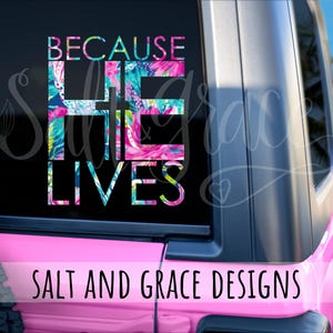 May include: A pink vehicle window decal with the words "BECAUSE HE LIVES" in a colorful floral pattern. The text "SALT AND GRACE DESIGNS" is below the main text. The decal is on a pink vehicle.