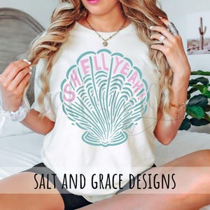 Puede incluir: Una camiseta blanca con un gr&aacute;fico de concha de mar rosa y azul y el texto "Shell Yeah!" impreso en la parte delantera. La camiseta tambi&eacute;n tiene el texto "Salt and Grace Designs" impreso en la parte inferior.