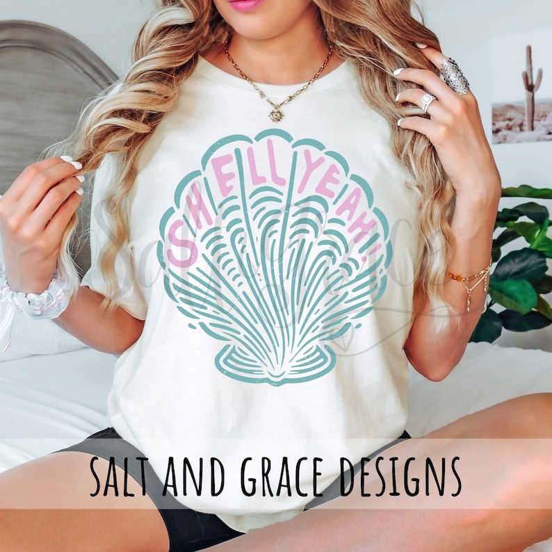 Puede incluir: Una camiseta blanca con un gr&aacute;fico de concha de mar rosa y azul y el texto "Shell Yeah!" impreso en la parte delantera. La camiseta tambi&eacute;n tiene el texto "Salt and Grace Designs" impreso en la parte inferior.