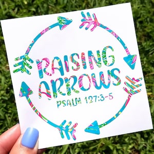 May include: White sticker with a floral design in shades of pink, blue, and green. The sticker features a circle with arrows pointing in different directions. The text "Raising Arrows" is written in a bold font inside the circle. The text "Psalm 127:3-5" is written below the circle.