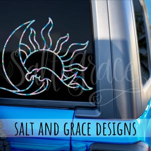 Op de afbeelding: Een decoratieve autosticker met een zon- en maanontwerp met een bloemenpatroon in blauwe, roze en groene tinten. De sticker is aangebracht op een autoraam. De tekst "SALT AND GRACE DESIGNS" staat onderaan.