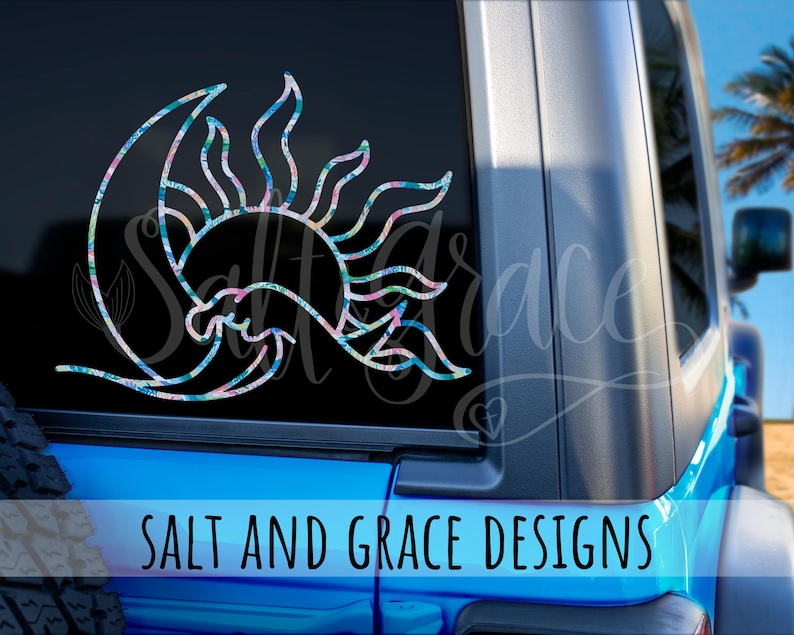 Op de afbeelding: Een decoratieve autosticker met een zon- en maanontwerp met een bloemenpatroon in blauwe, roze en groene tinten. De sticker is aangebracht op een autoraam. De tekst "SALT AND GRACE DESIGNS" staat onderaan.