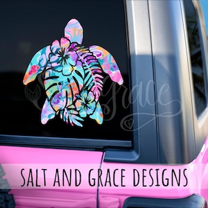 May include: A colorful sea turtle decal with a floral pattern in shades of blue, pink, orange, and purple is displayed on a vehicle window. The decal features the text "SALT AND GRACE DESIGNS" in white. The background shows a glimpse of a sunny day.